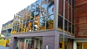 Un edificio moderno con amplios ventanales que reflejan palmeras, un área de estar exterior cubierta con cojines, y una fachada púrpura.
