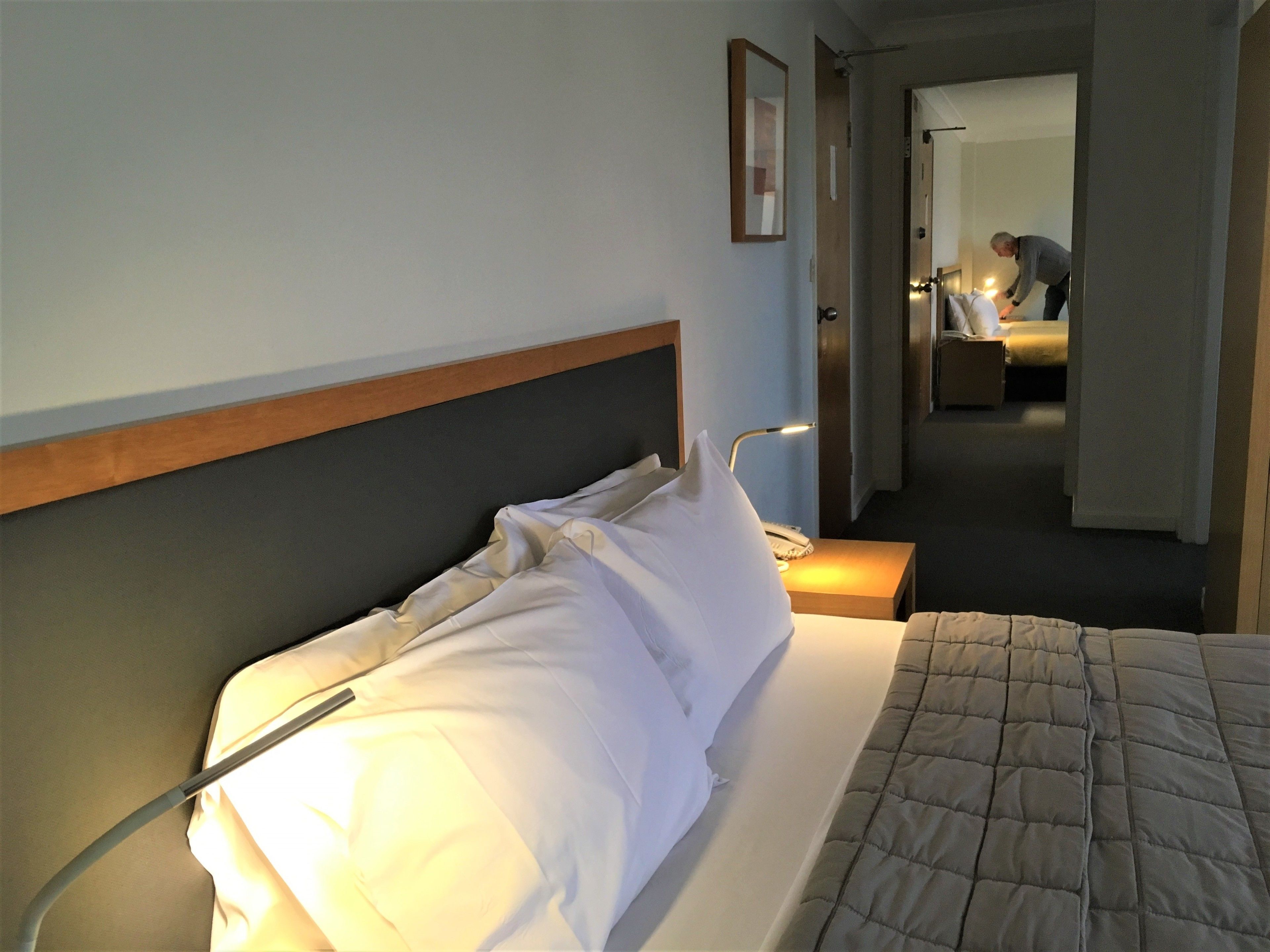 Ett hotellrum med en säng, ett nattduksbord, en lampa och en dörr som leder till ett annat rum.