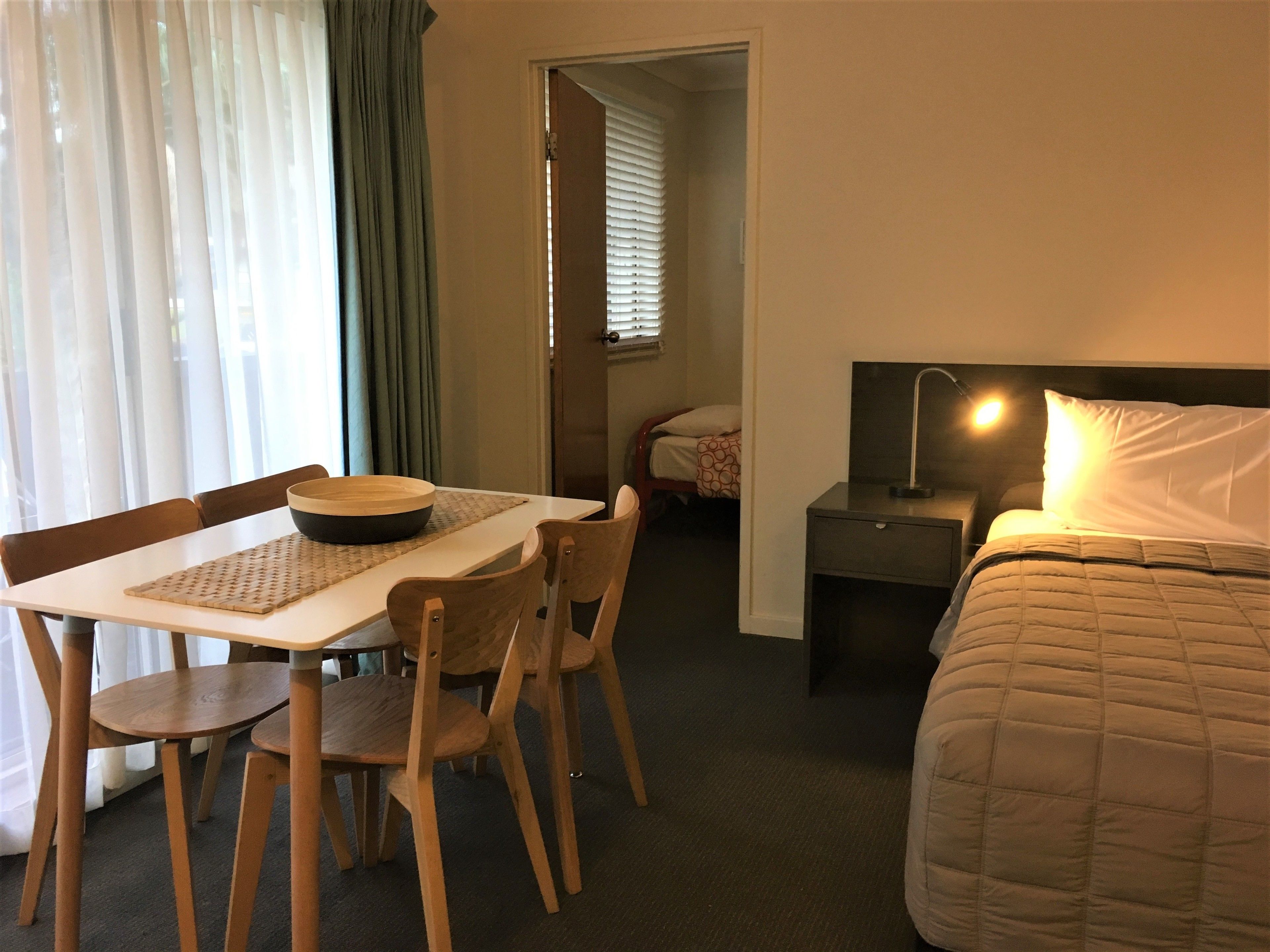 Habitación de hotel con cama, mesita de noche, lámpara y un área de comedor con mesa y sillas.