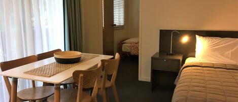 Habitación de hotel con cama, mesita de noche, lámpara y un área de comedor con mesa y sillas.