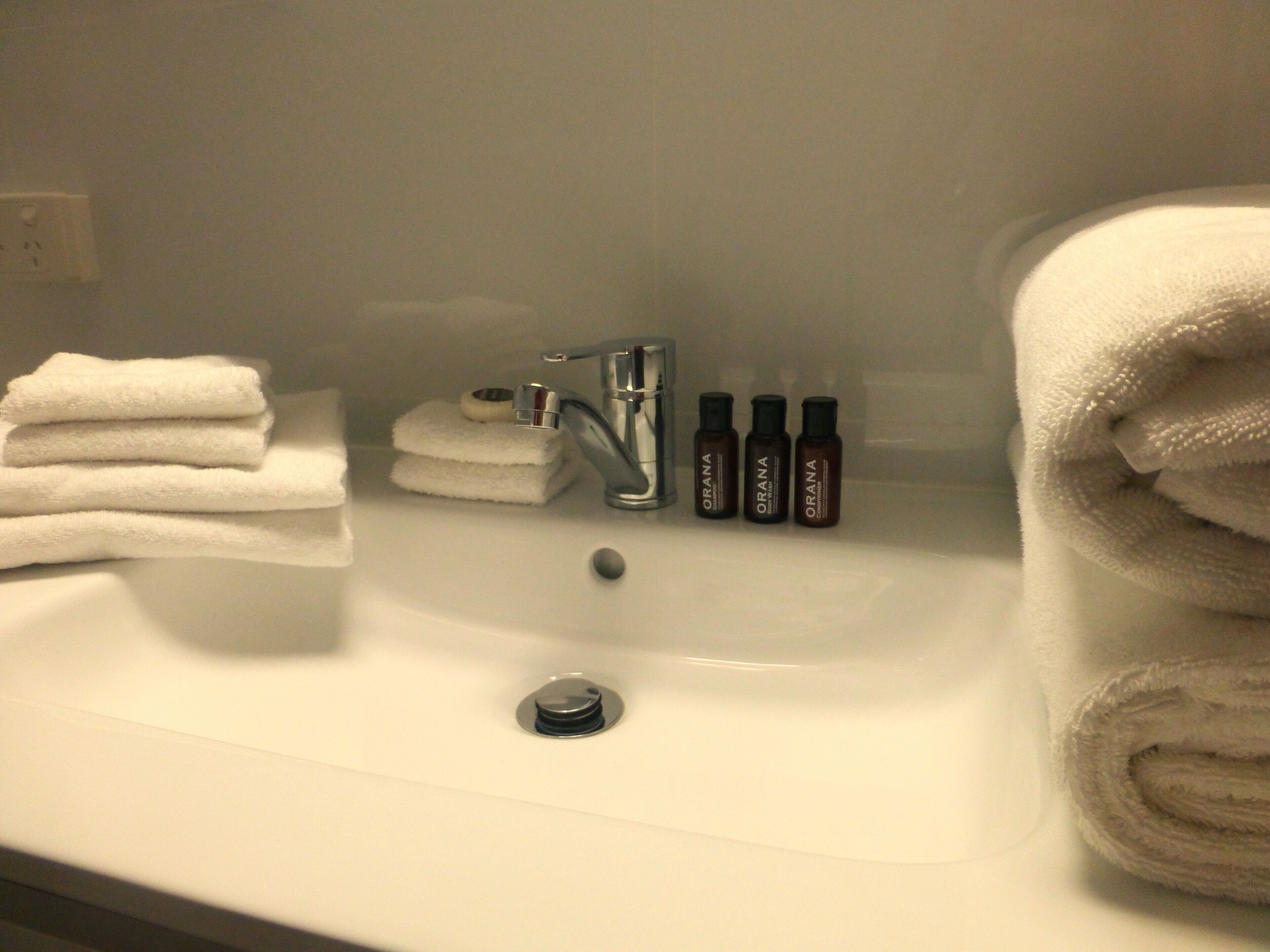 A bathroom sink with stacked white towels, a faucet, and three bottles labeled "ORANA".