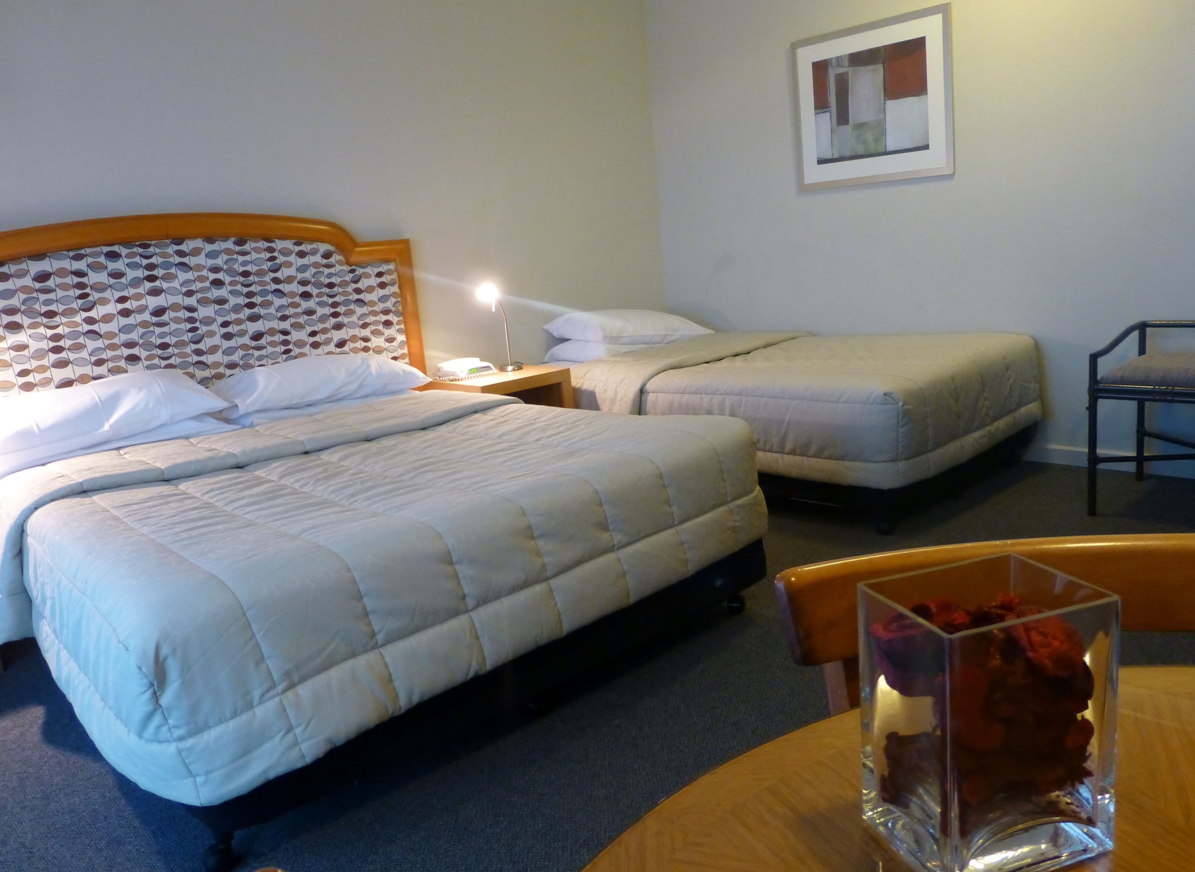 Habitación de hotel con dos camas, cabecero de madera, mesita de noche con lámpara, una silla y un jarrón con flores.