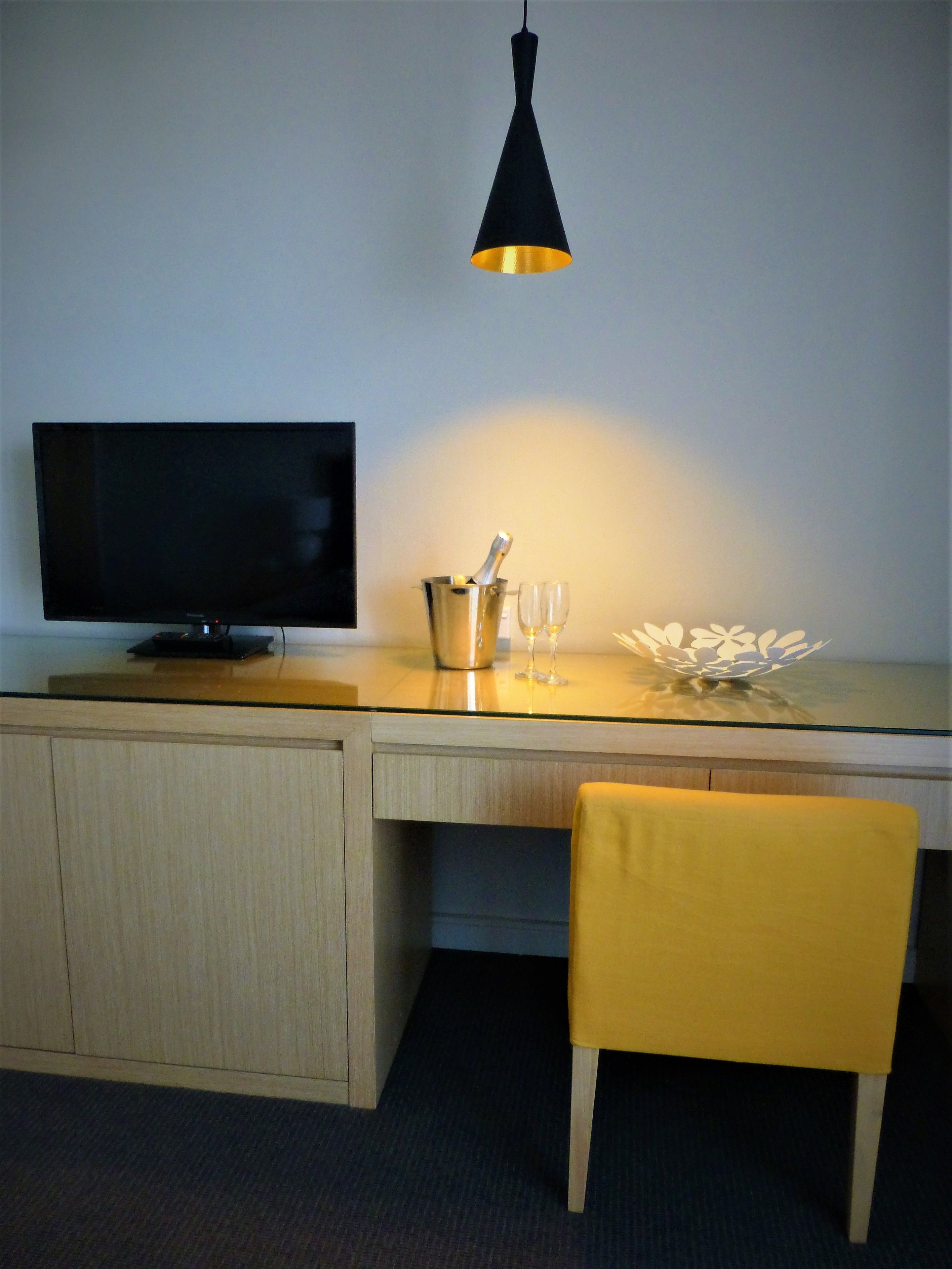 A modern room with a wooden desk, a television, a yellow chair, a black pendant light, and a decorative object on the desk.