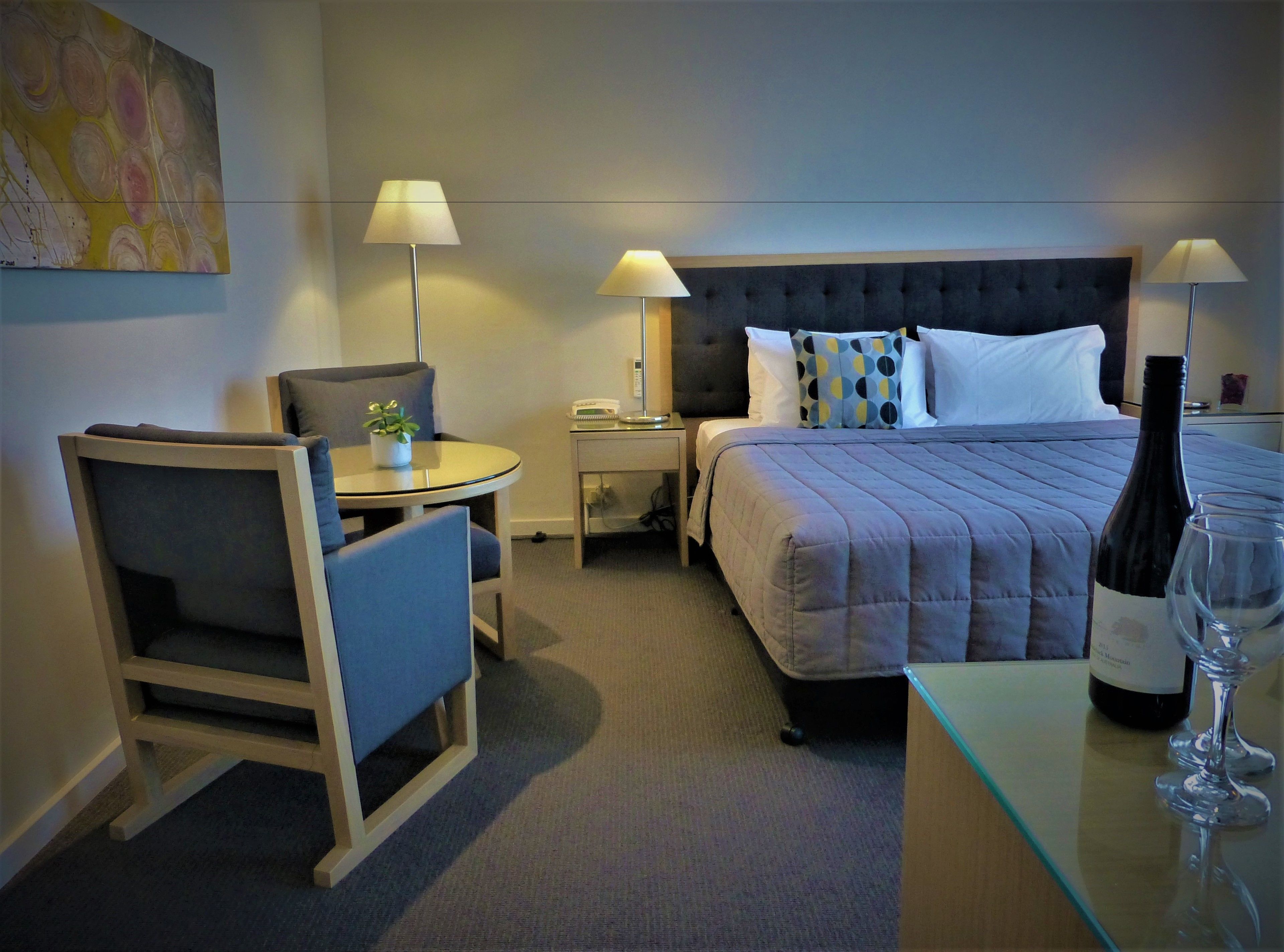 A hotel room with a bed, bedside tables, a desk with a chair, a lamp, a bottle of wine, and a glass.