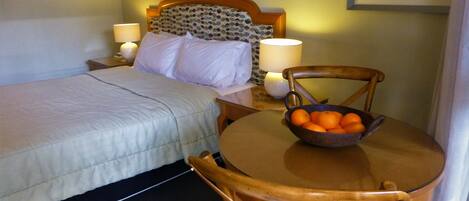 A hotel room with a bed, bedside lamps, a small table with a fruit bowl, and a chair.