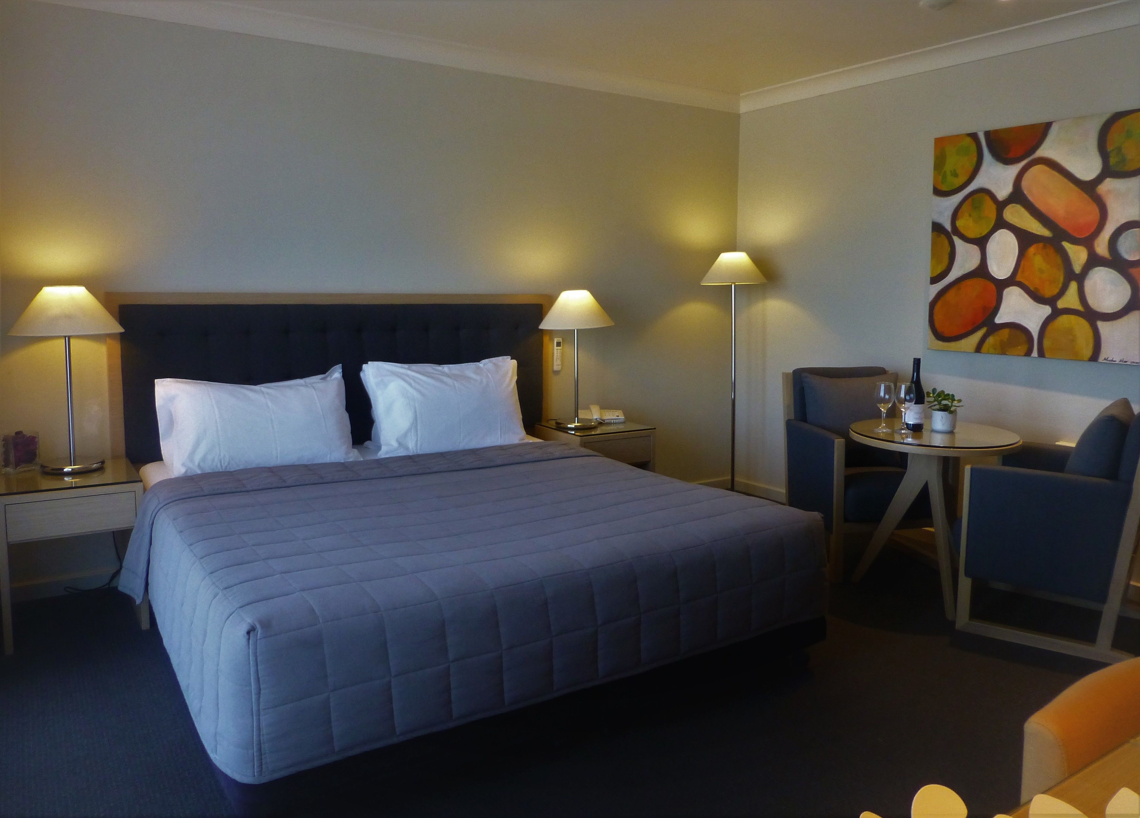 A hotel room with a large bed, two bedside lamps, a small table with a chair, and a painting on the wall.