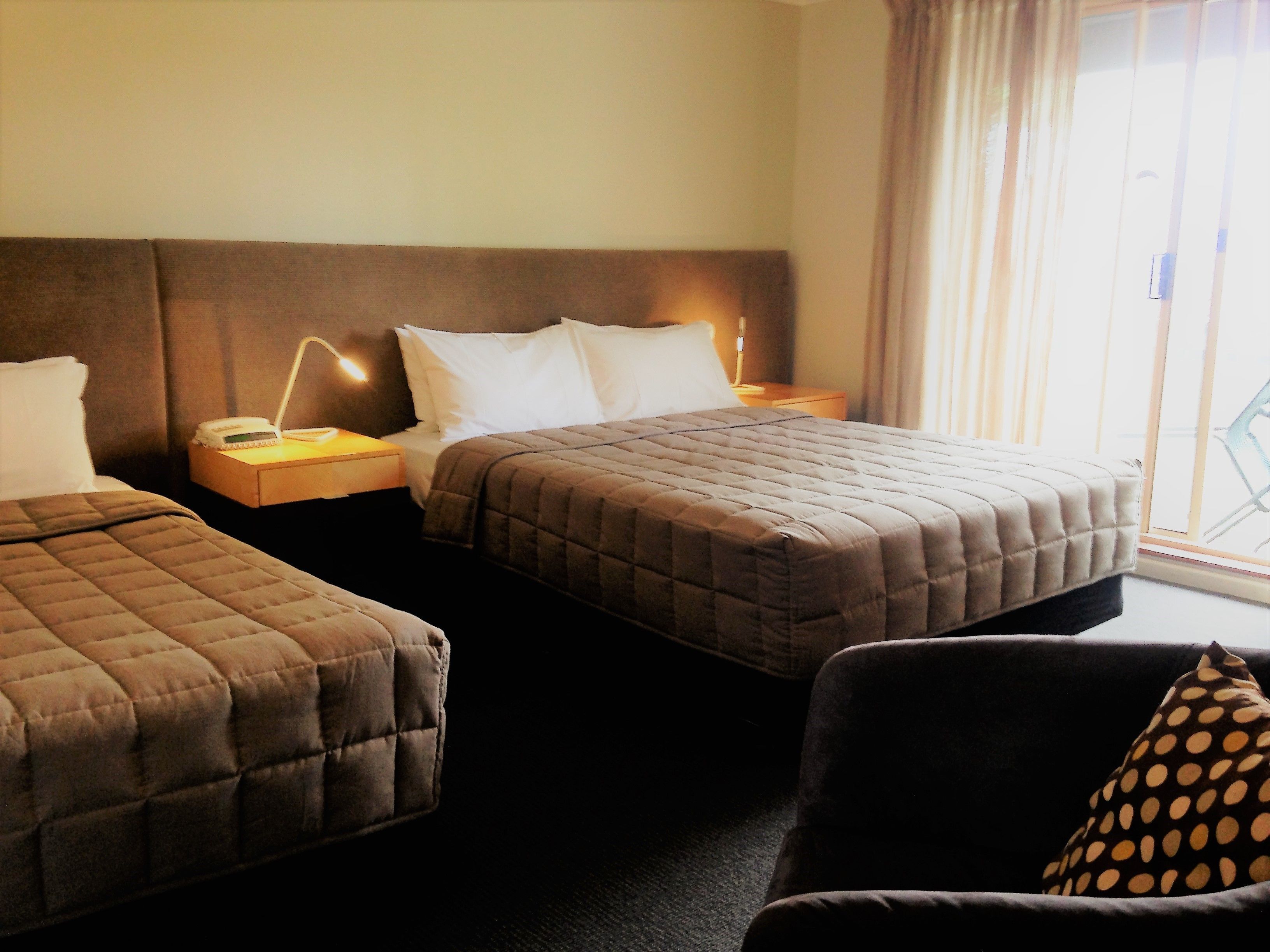 A hotel room with two beds, a nightstand with a lamp, and a view of the outdoors through a window.