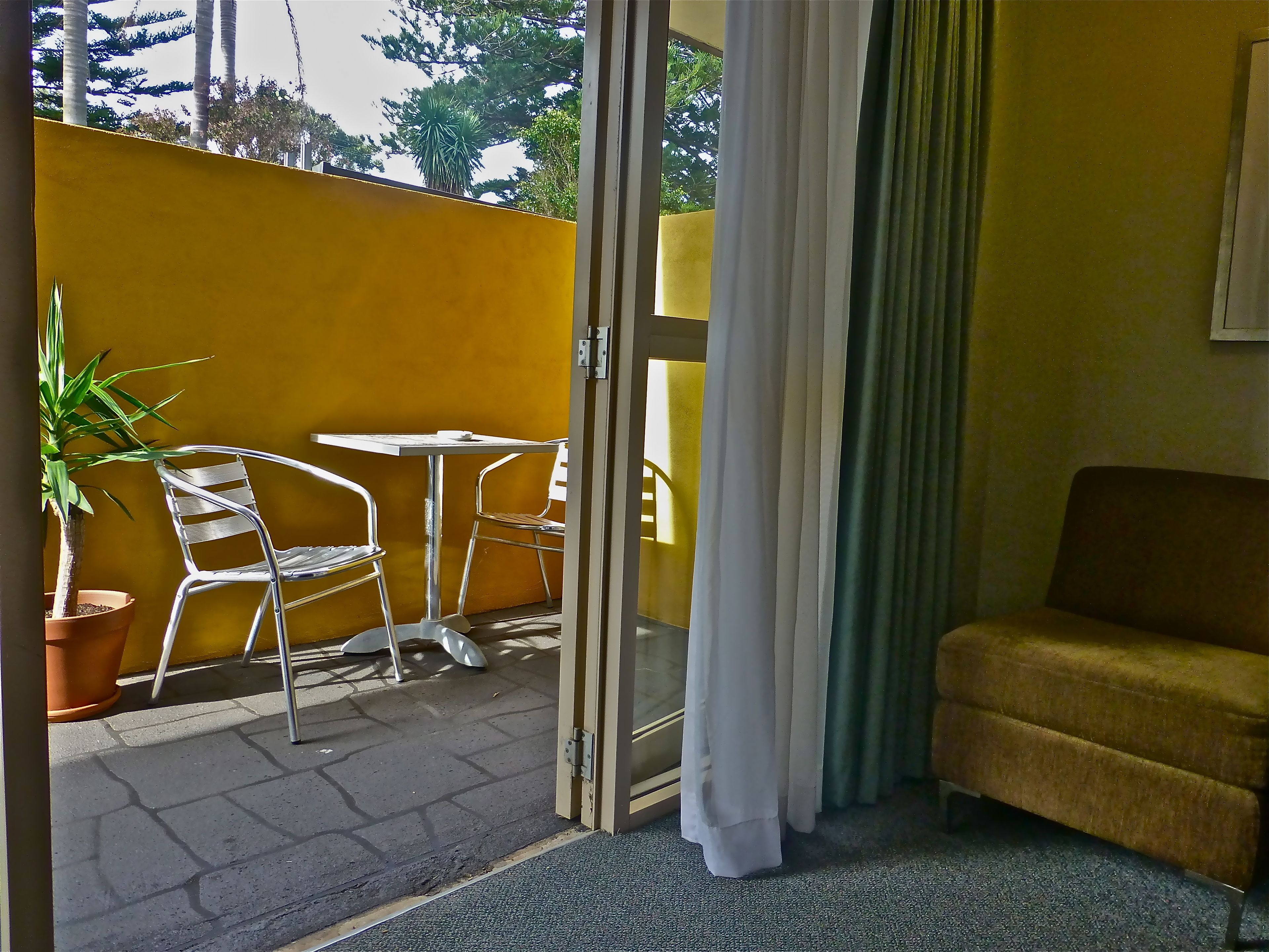 Habitación con puerta de vidrio corredera que da a un espacio exterior con pared amarilla, planta en maceta, dos sillas de metal y una pequeña mesa.