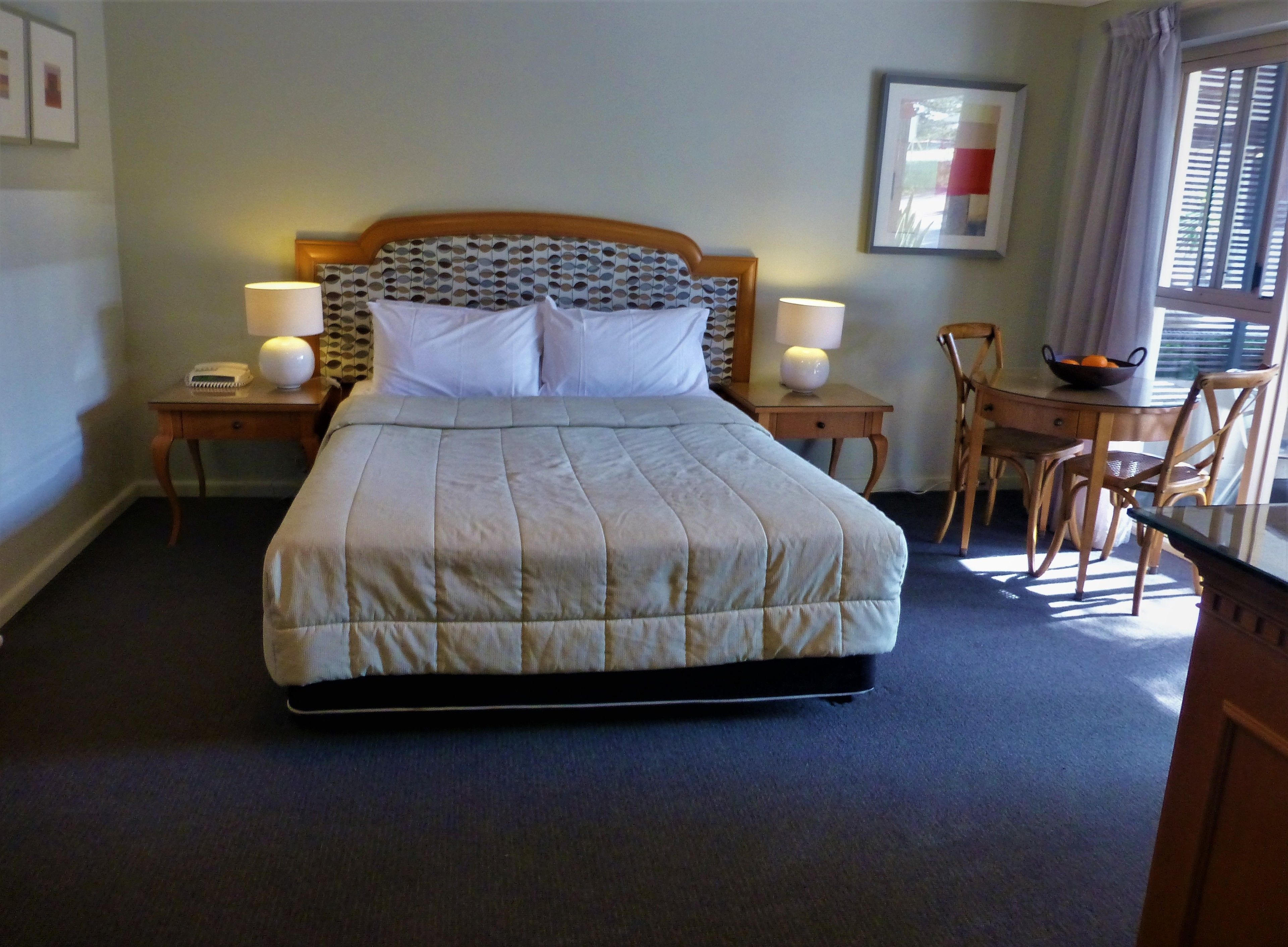A neatly arranged hotel room with a large bed, two bedside tables with lamps, a wooden chair, and a small table with a bowl of fruit.