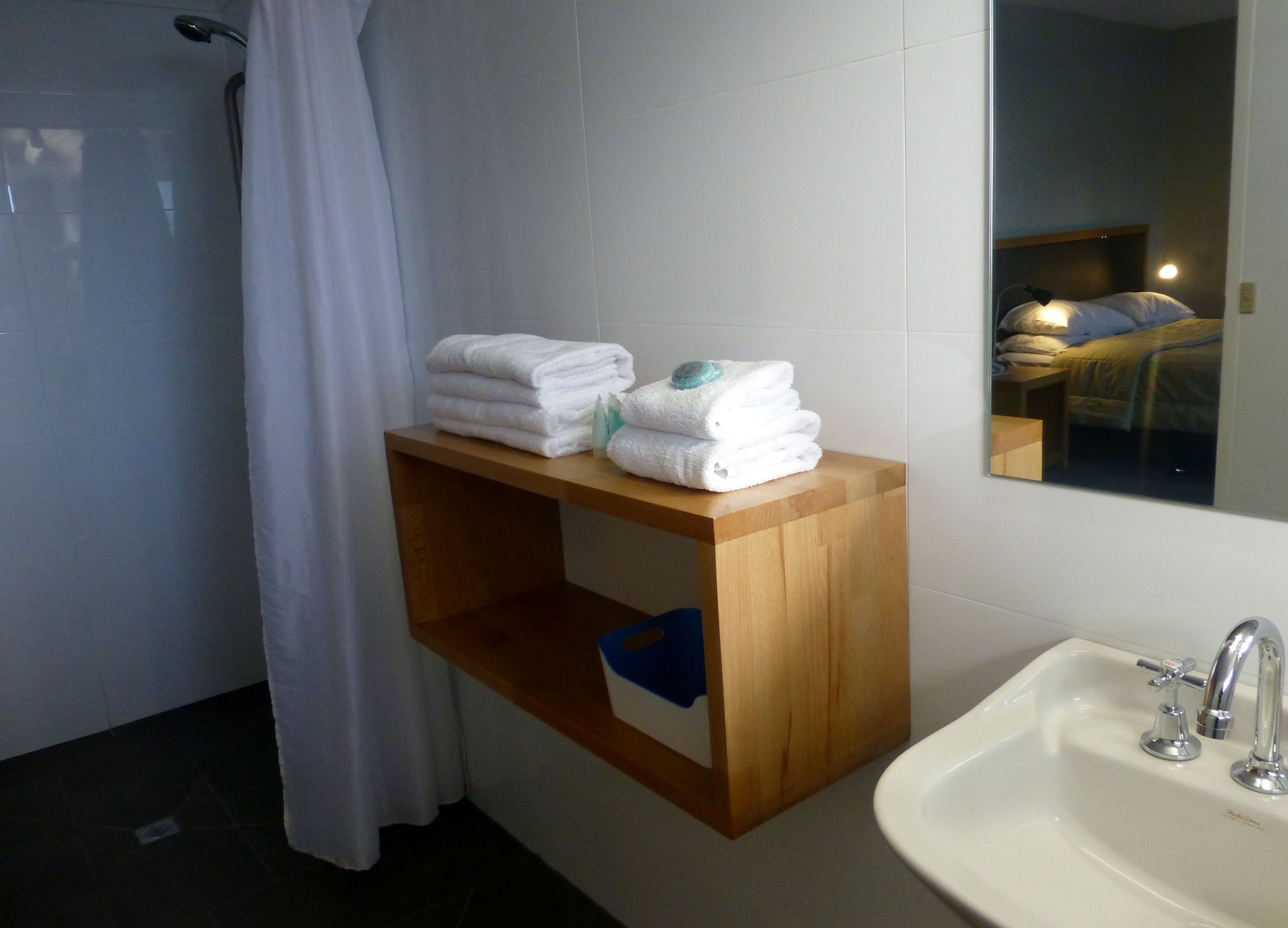 A bathroom with a wooden shelf holding folded towels, a sink with a mirror, and a shower area.