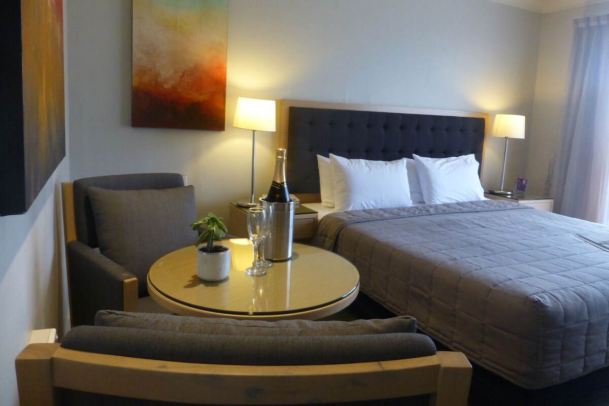 A hotel room with a bed, two bedside tables, a round table with a plant, a chair, and a painting on the wall.