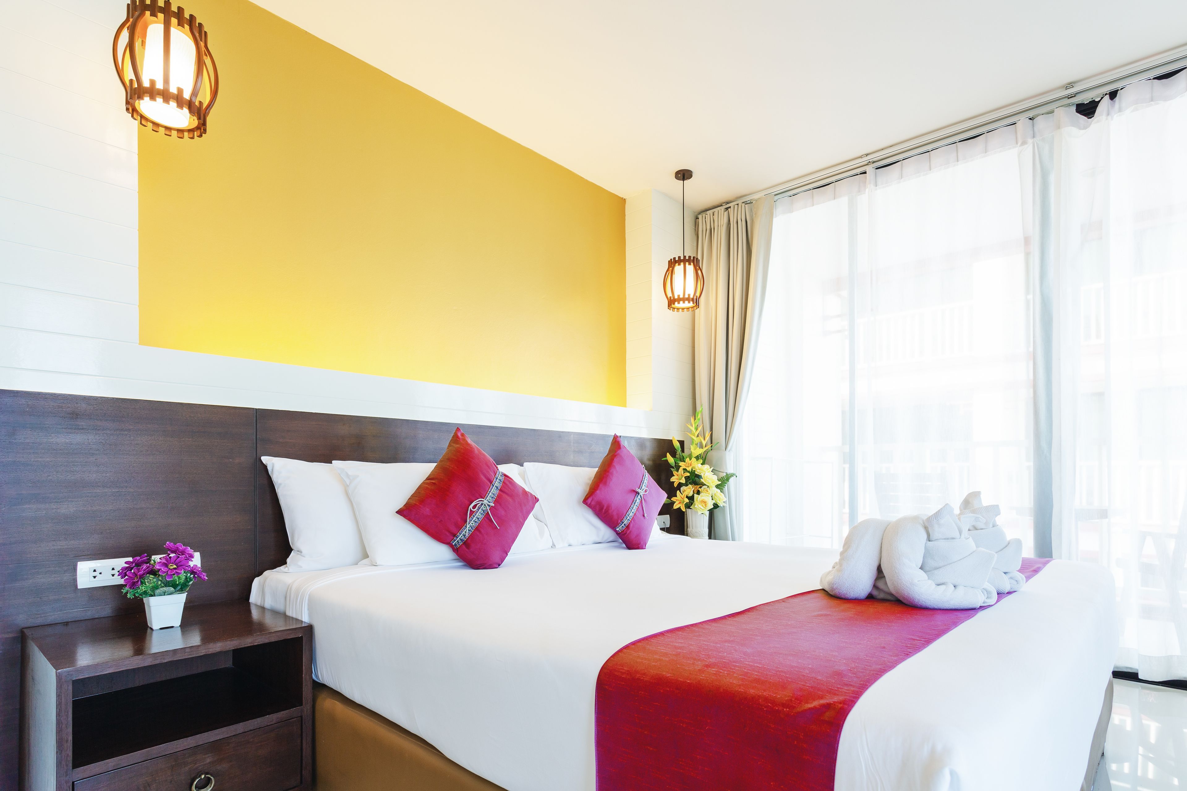 A modern hotel room with a large bed, a nightstand with a vase of flowers, and a bright yellow accent wall.