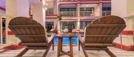 A poolside area with two wooden rocking chairs and a table, a pool, and a multi-level building with balconies in the background.