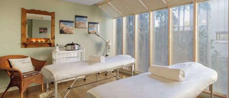 A room with two beds, a wicker chair, a mirror, and a window with blinds.