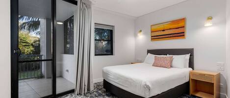 A bedroom with a large bed, a sliding glass door to a balcony, a ceiling fan, and a painting on the wall.