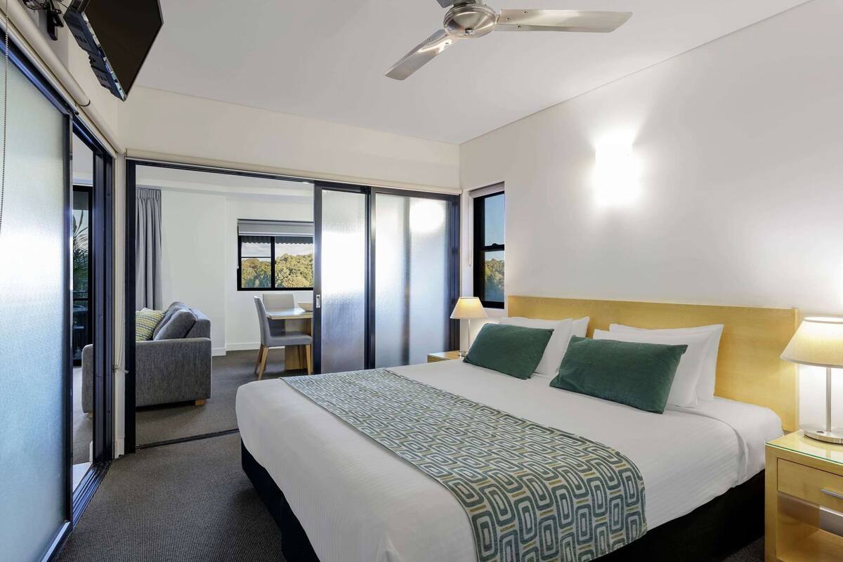 A hotel room with a bed, a desk, a chair, a ceiling fan, and a view of the outdoors.