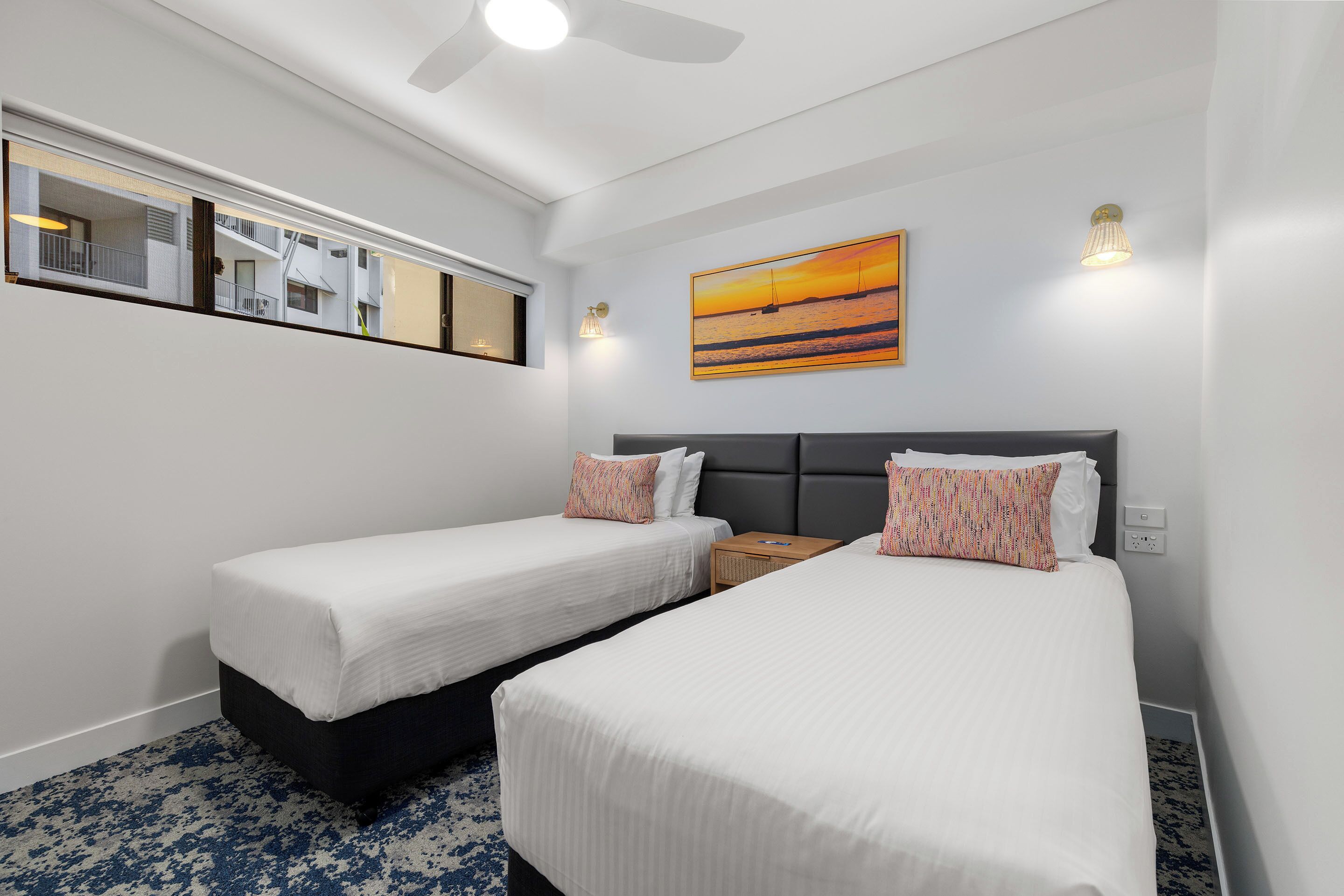 A hotel room with two single beds, a ceiling fan, a painting of a sunset, and a window with a view of another building.
