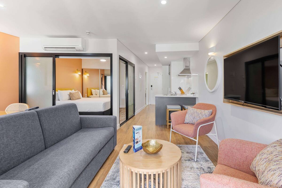 A modern living room with a grey sofa, a pink armchair, and a round coffee table. There is a TV mounted on the wall, a small table with a bowl and a remote control, and a sliding door leading to a bedroom.
