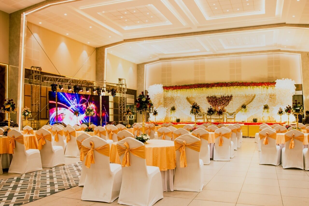 Un salón de banquetes con mesas redondas, sillas blancas con cintas naranjas y un escenario con decoración floral.