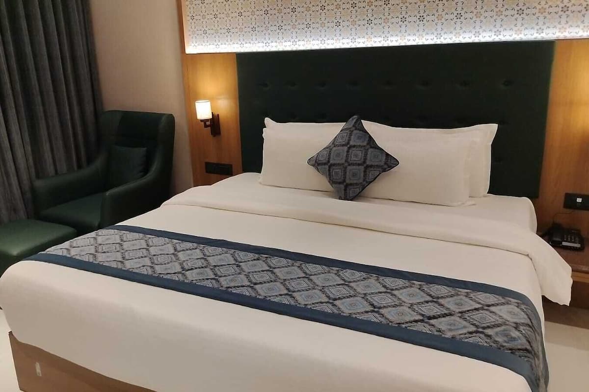 A hotel room with a bed, a green armchair, and a nightstand with a lamp.