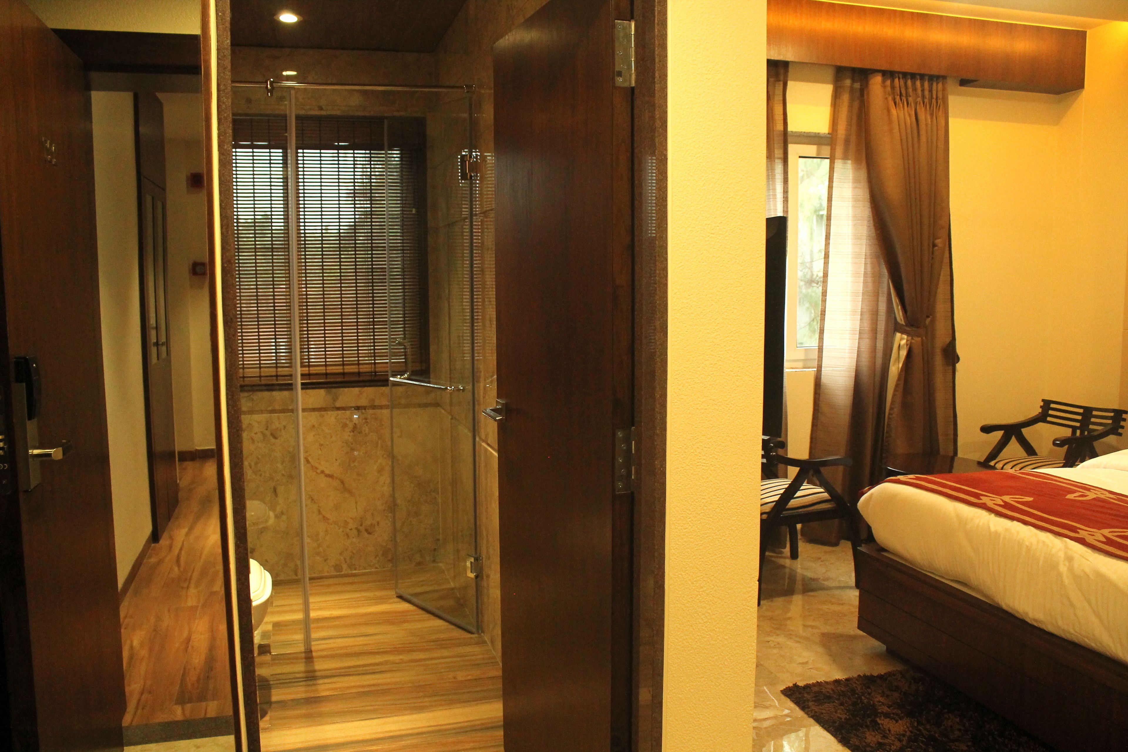 Habitación de hotel con cabina de ducha de vidrio, suelo de madera, cama con edredón rojo y blanco, y una silla de madera.