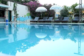 A swimming pool with lounge chairs and umbrellas, surrounded by a wall with murals and plants. - Hotel Surya (Varanasi)