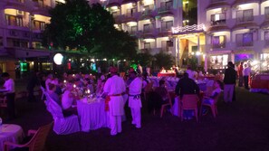 A formal outdoor dining area with round tables, guests seated, and waitstaff in uniform. - Hotel Surya (Varanasi)