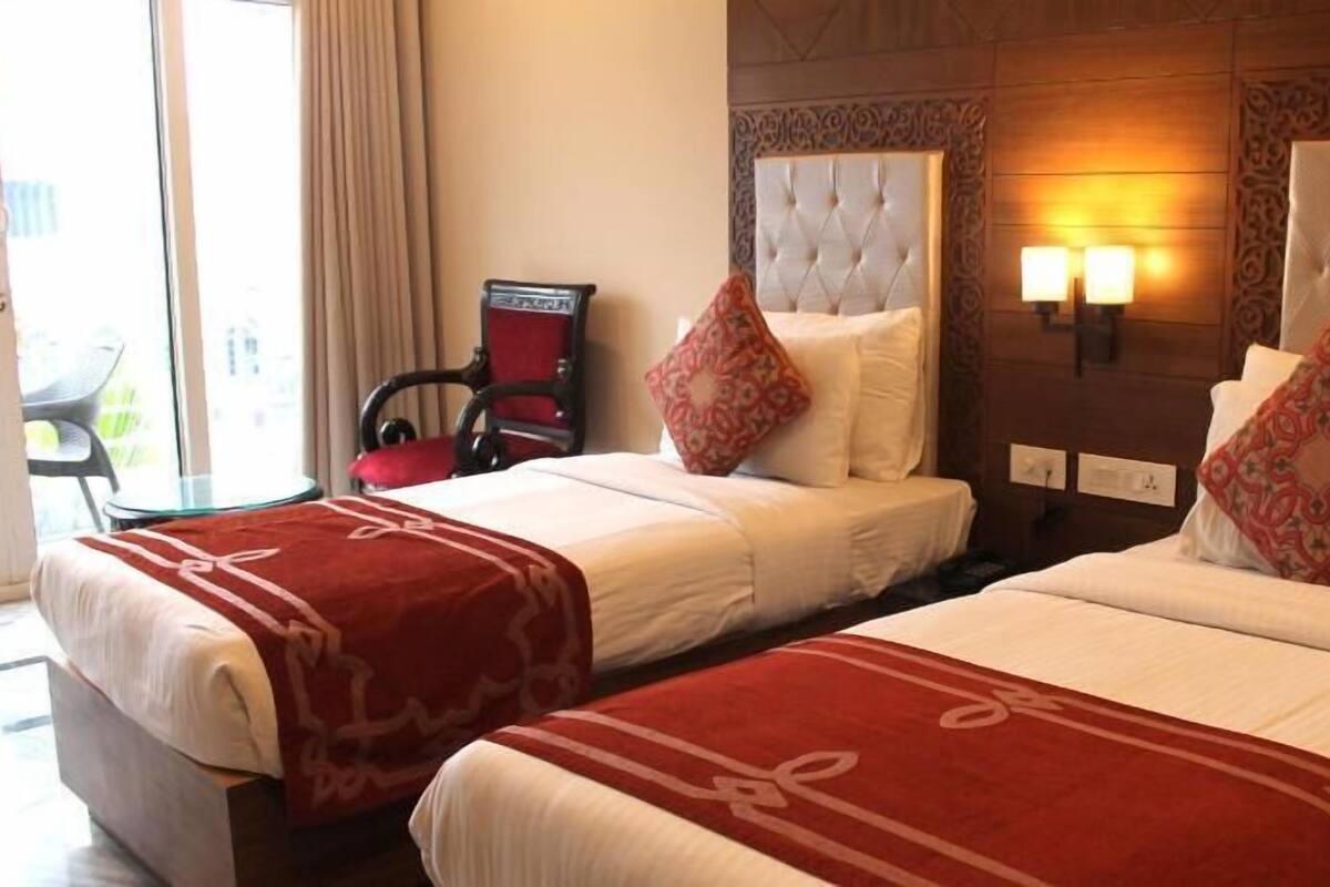 A hotel room with two beds, a red and white bedspread, a red chair, a wooden headboard, and a wall-mounted light fixture.
