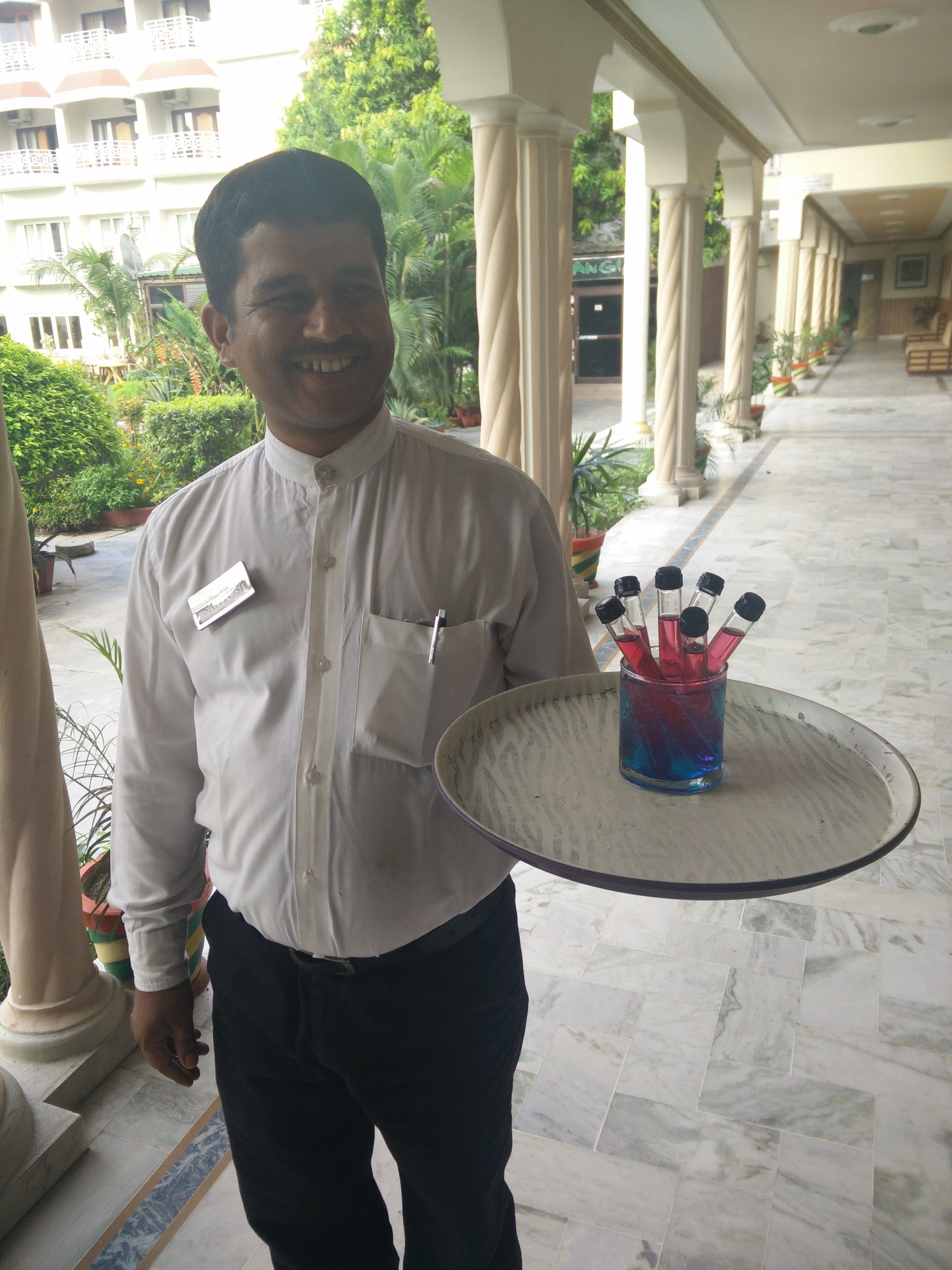 A man in a white shirt holding a tray with a glass and utensils, standing in a corridor with columns and potted plants.