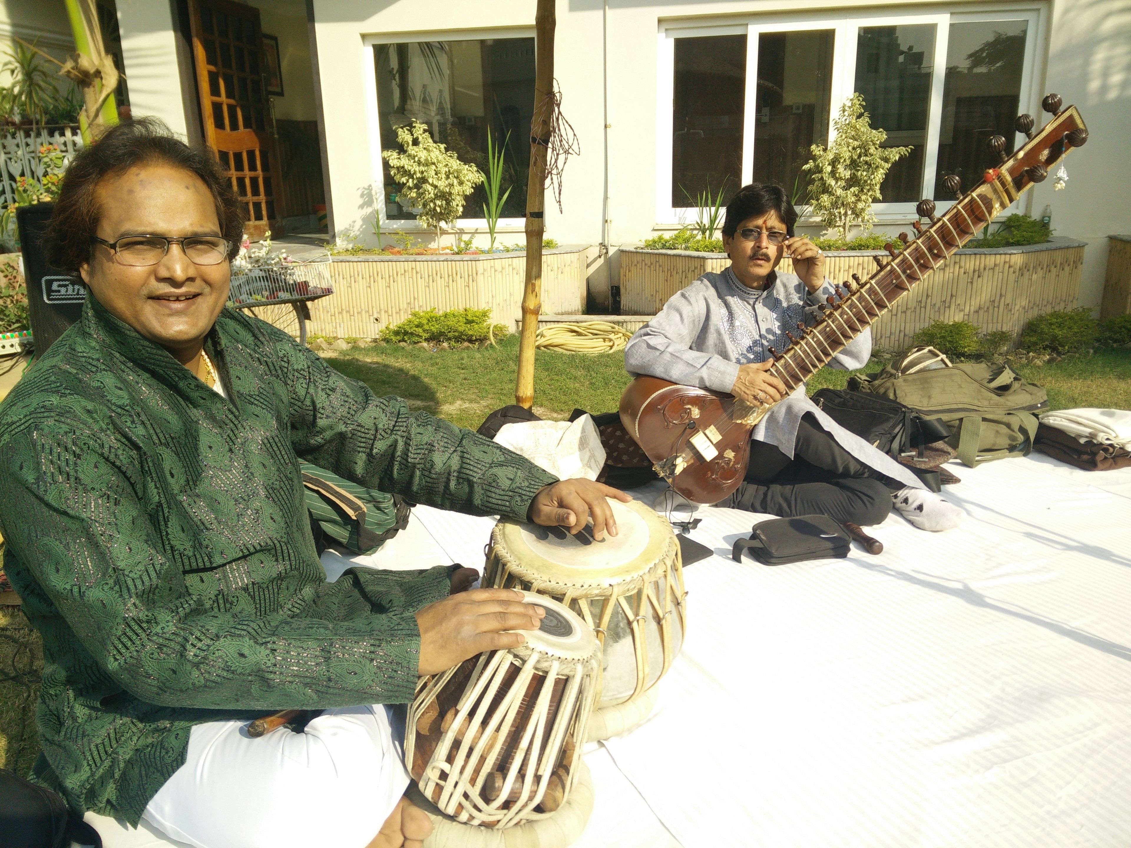 Two musicians seated on a white outdoor mat, playing traditional string and percussion instruments.