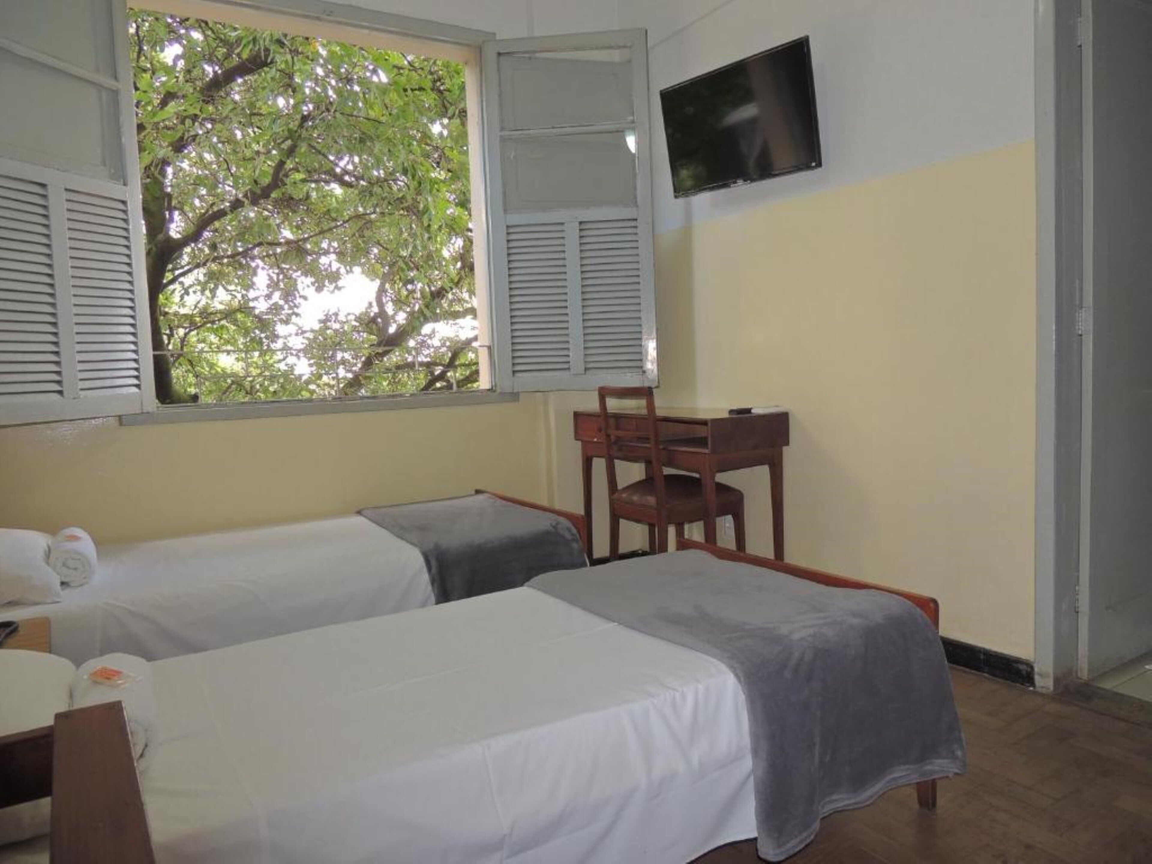 A room with two beds, a desk, a chair, a TV, and a window with shutters.
