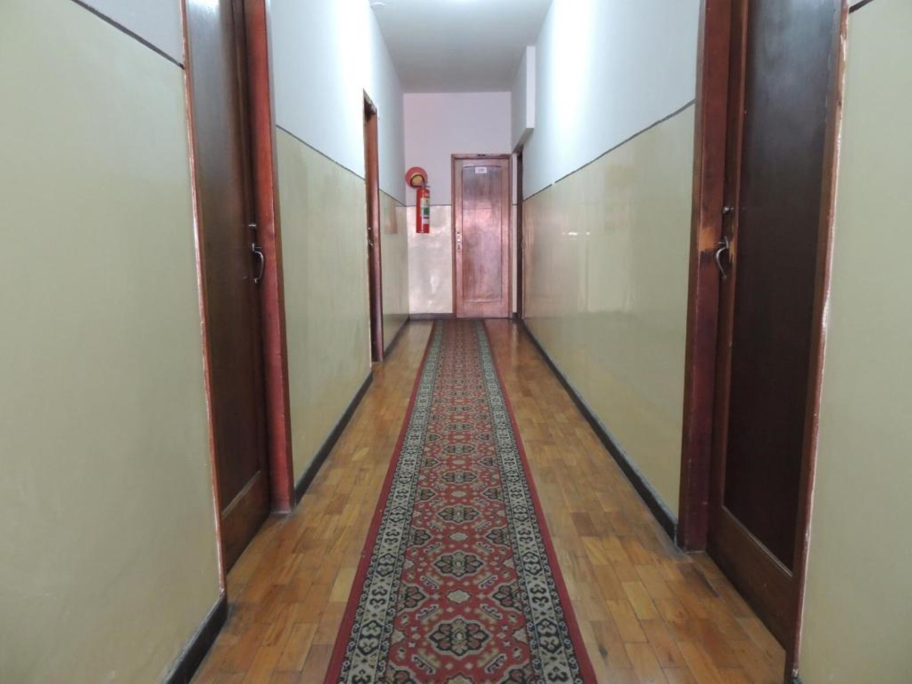 Un pasillo con puertas de madera, alfombra estampada y un extintor en la pared.