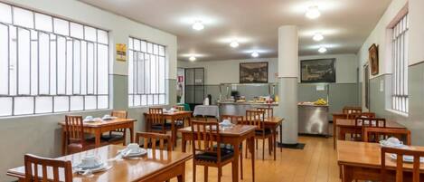Un espace repas avec des tables et des chaises en bois, une cuisine à l’arrière-plan et de grandes fenêtres avec des barreaux.