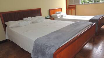 Cama de casal com cabeceira de madeira, manta cinza e lençóis brancos. Há um banquinho de madeira e uma pequena mesa com um telefone verde e um objeto branco.