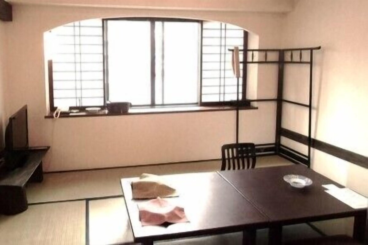 A room with tatami flooring, a low table with a cushion, a chair, a shelf with a TV, and a window with blinds.