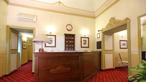 Zona de recepción de hotel con mostrador de madera, un reloj en la pared, cuadros enmarcados y piso cubierto de alfombra roja.
