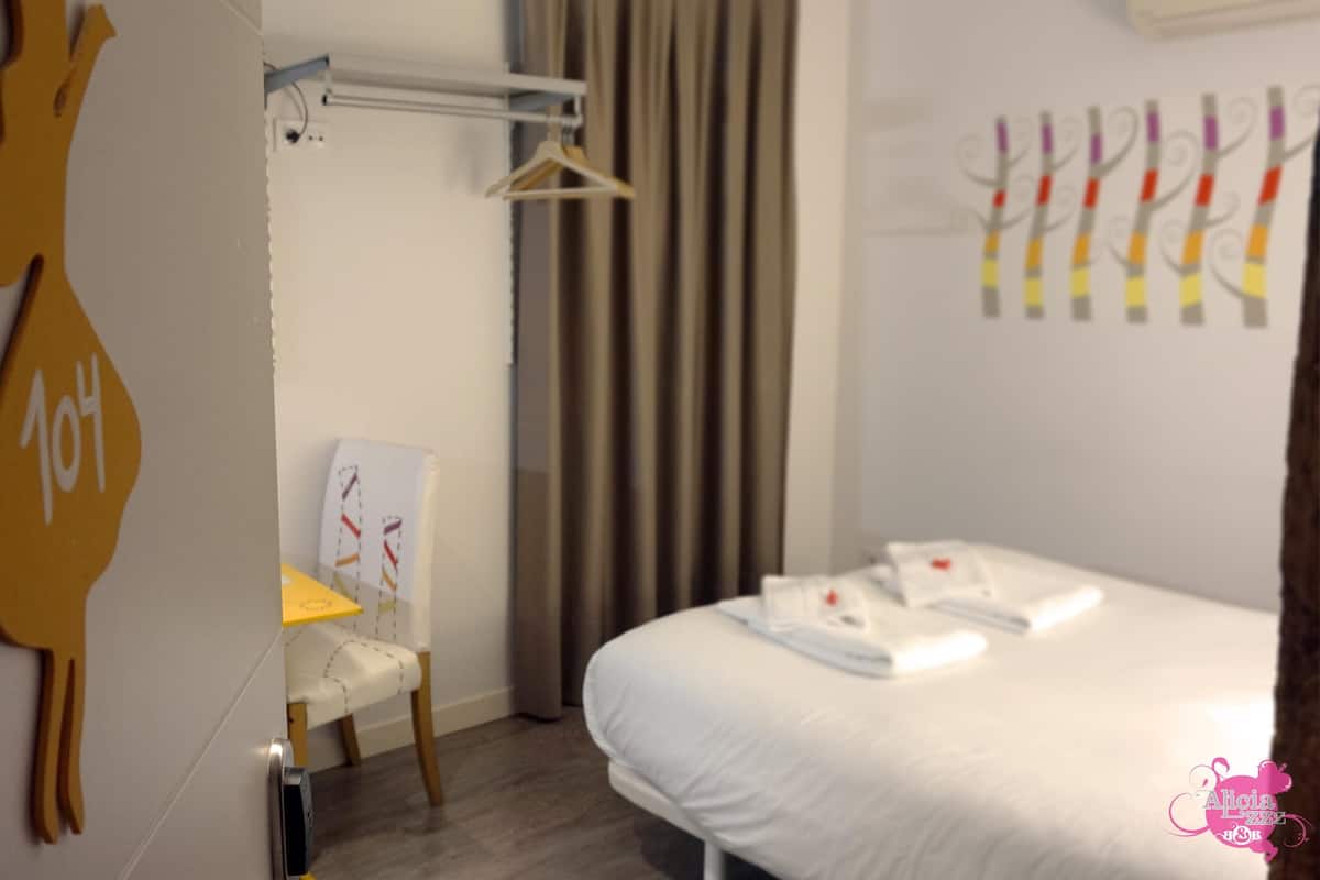 Habitación de hotel con cama, escritorio con silla, perchero y un armario con ropa colgada.