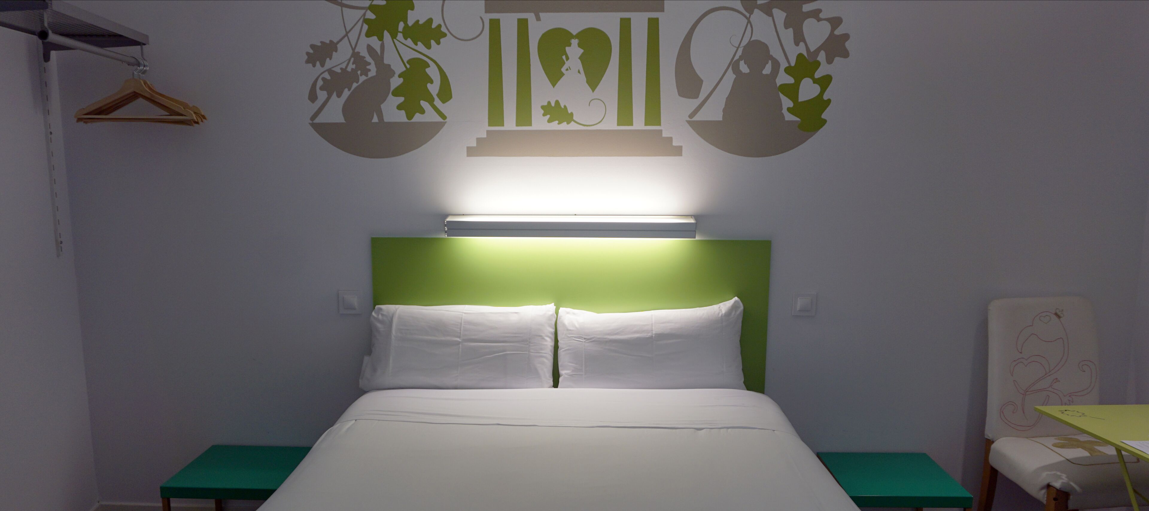 Een opgemaakt bed met witte lakens, een groen hoofdeinde en een nachtkastje met een groen oppervlak.