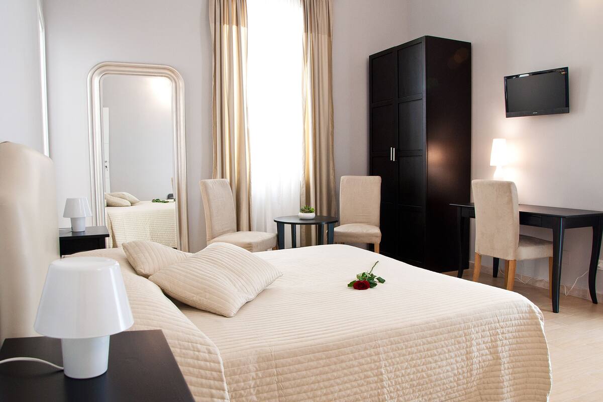 A hotel room with a bed, two chairs, a desk, a wardrobe, a mirror, and a TV.