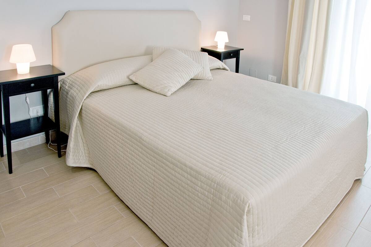 A neatly made bed with a quilted bedspread and two pillows, flanked by two bedside tables with lamps, in a room with light-colored flooring and curtains.