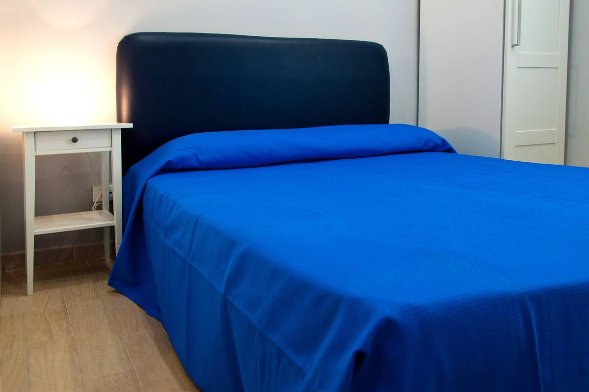 A single bed with a blue bedspread and a matching pillow, a white nightstand with a drawer, and a white door in the background.