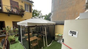 Un área de patio techada con una mesa y sillas, rodeada de plantas en macetas y un edificio amarillo al fondo.