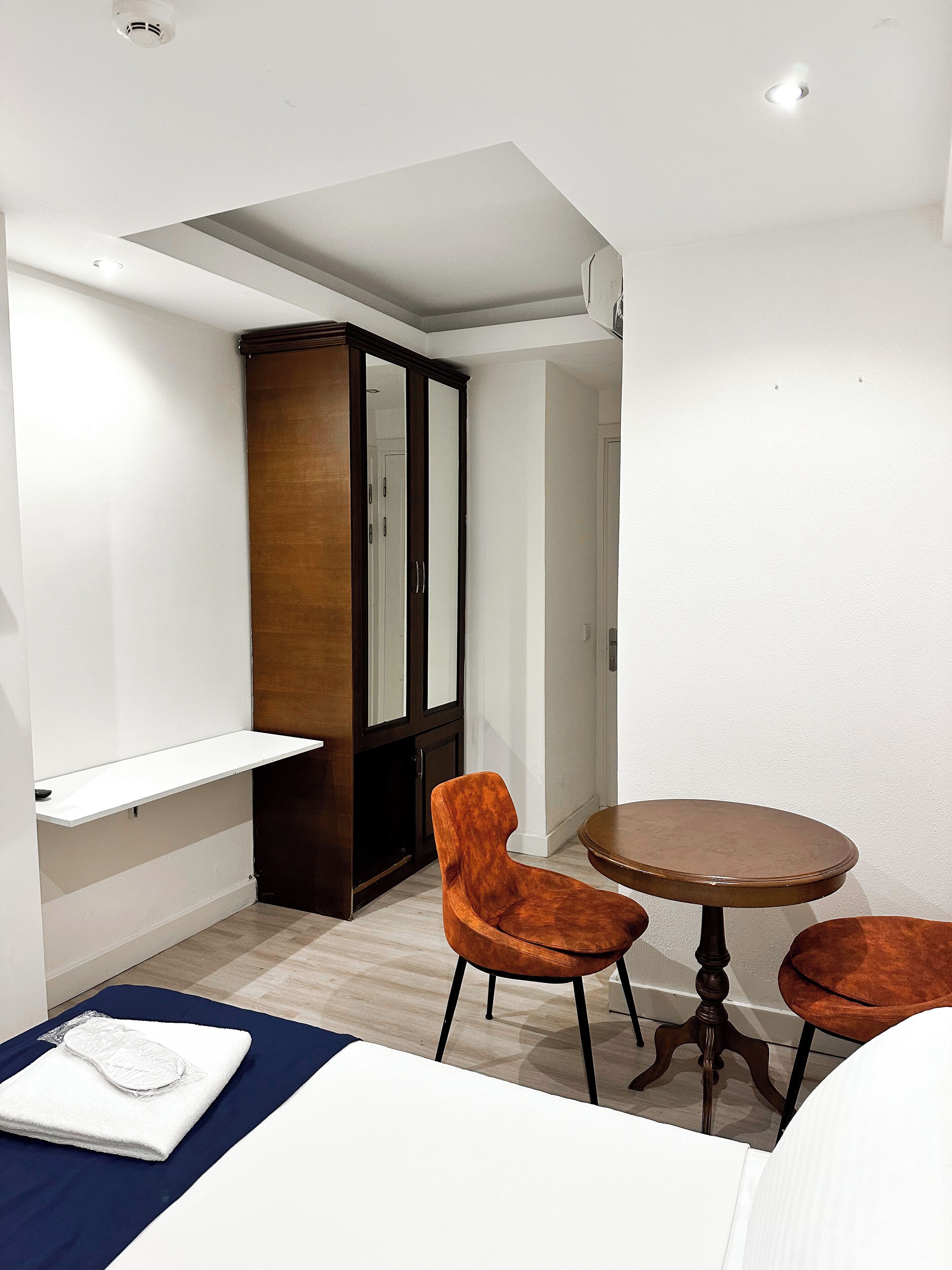 A compact room with a wooden wardrobe, two chairs, a small round table, and a white shelf.