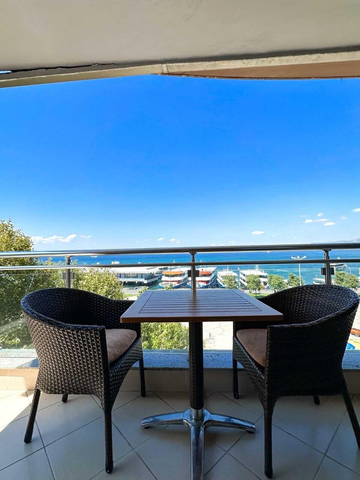 A balcony with a table and two wicker chairs, overlooking a coastal area with buildings and boats.