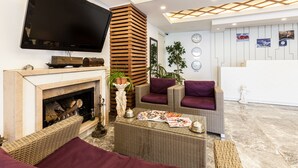 A modern living room with a flat-screen TV, a fireplace, wicker furniture, and a reception desk in the background. - Büyükada Comfort Hotel (Adalar)