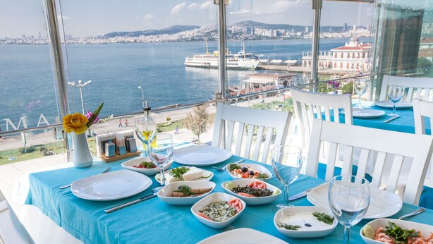 A dining table set with various dishes and glasses, overlooking a waterfront with a cityscape in the background. - Büyükada Comfort Hotel (Adalar)