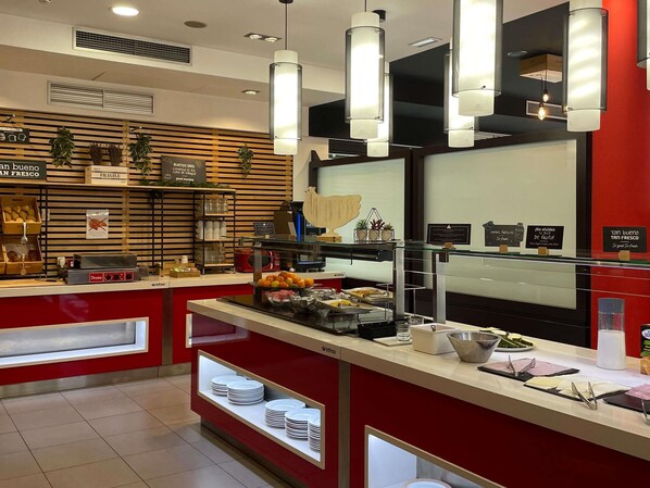 Una barra de desayuno moderna con una decoración roja y blanca, equipada con una cafetera, una exhibición de frutas y diversas opciones para el desayuno.