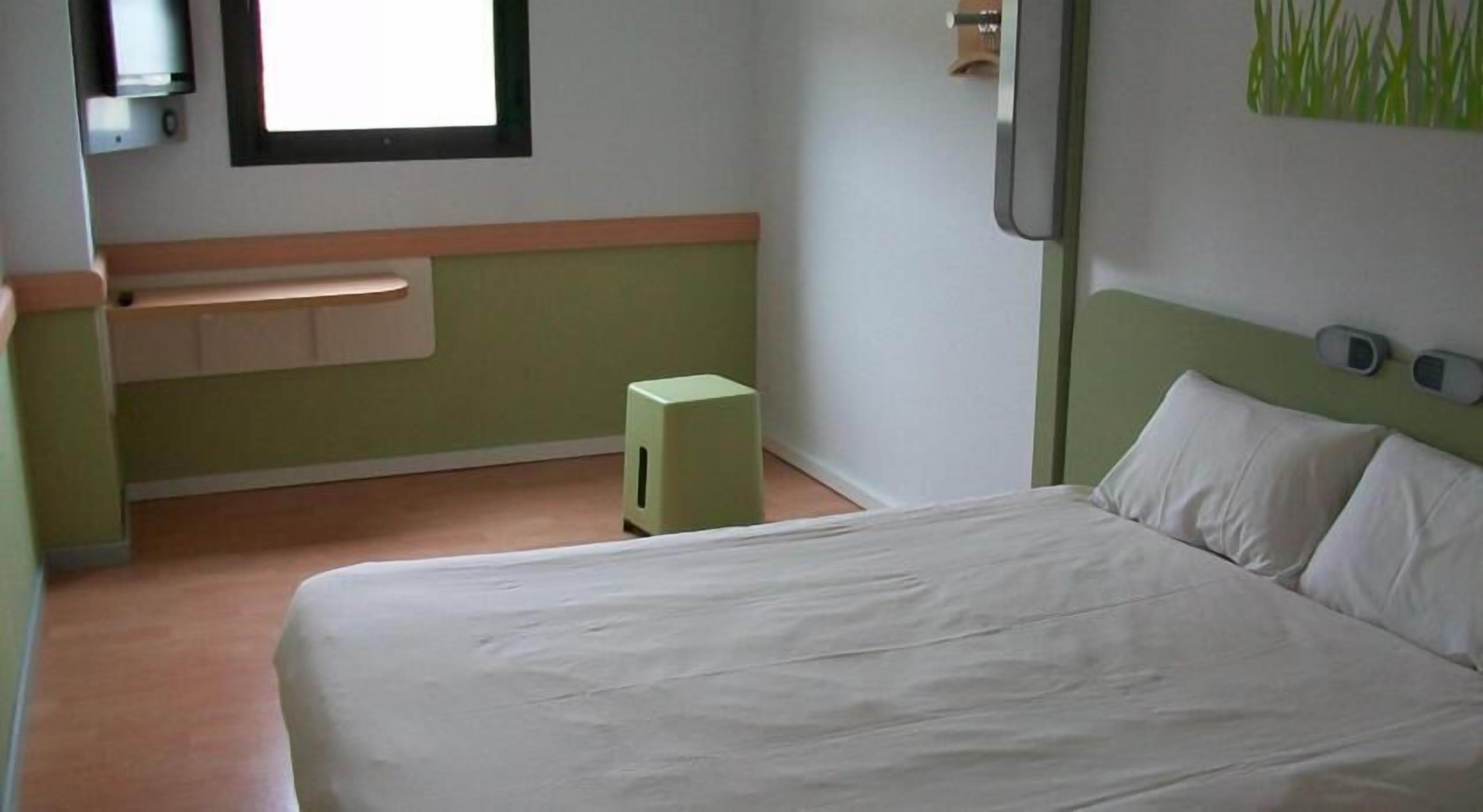 Une chambre d’hôtel avec un lit, un petit tabouret, une fenêtre et un luminaire fixé au mur.