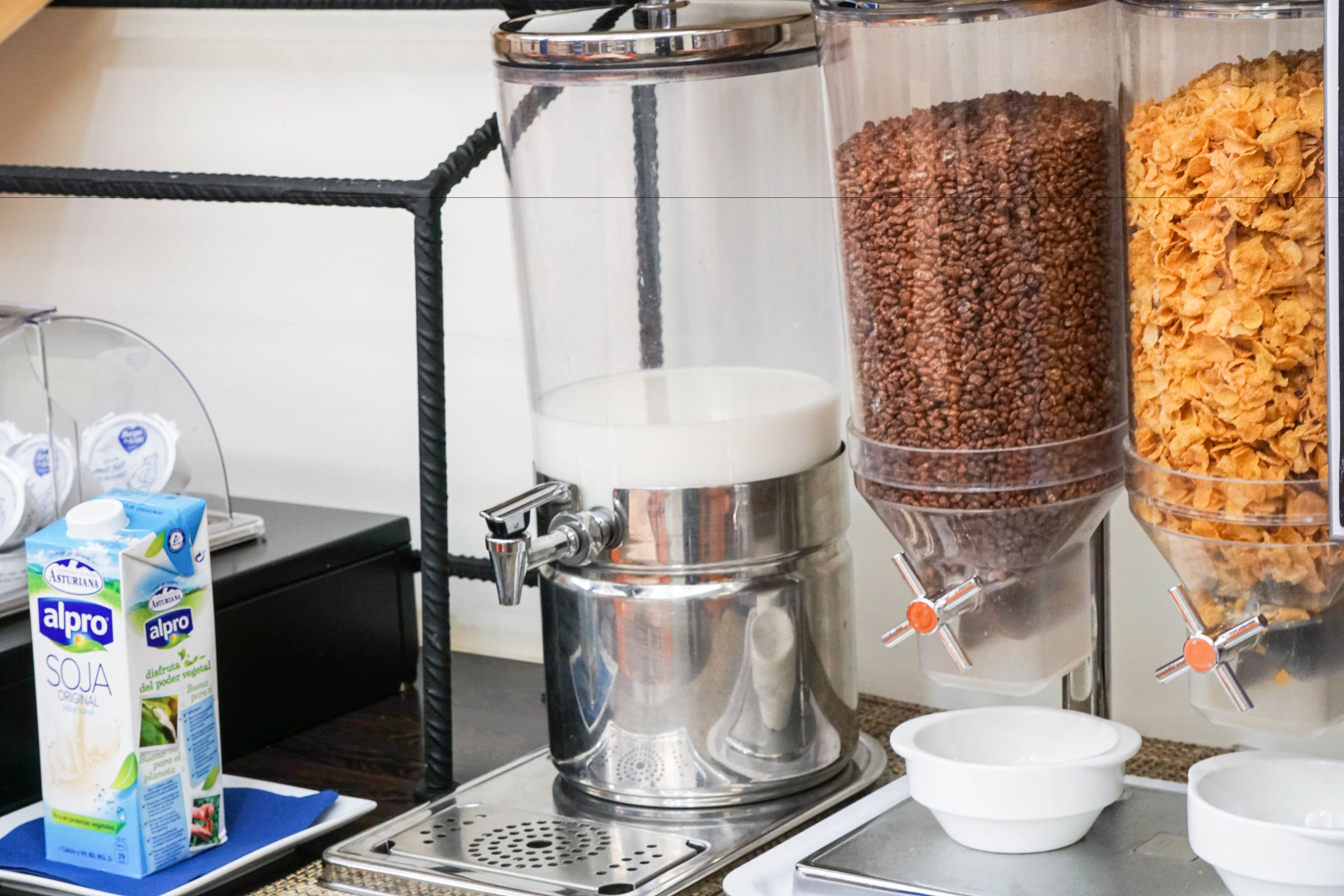 Una estación de café con dispensador de leche, máquina de café y recipientes con granos de café y copos de cereales.