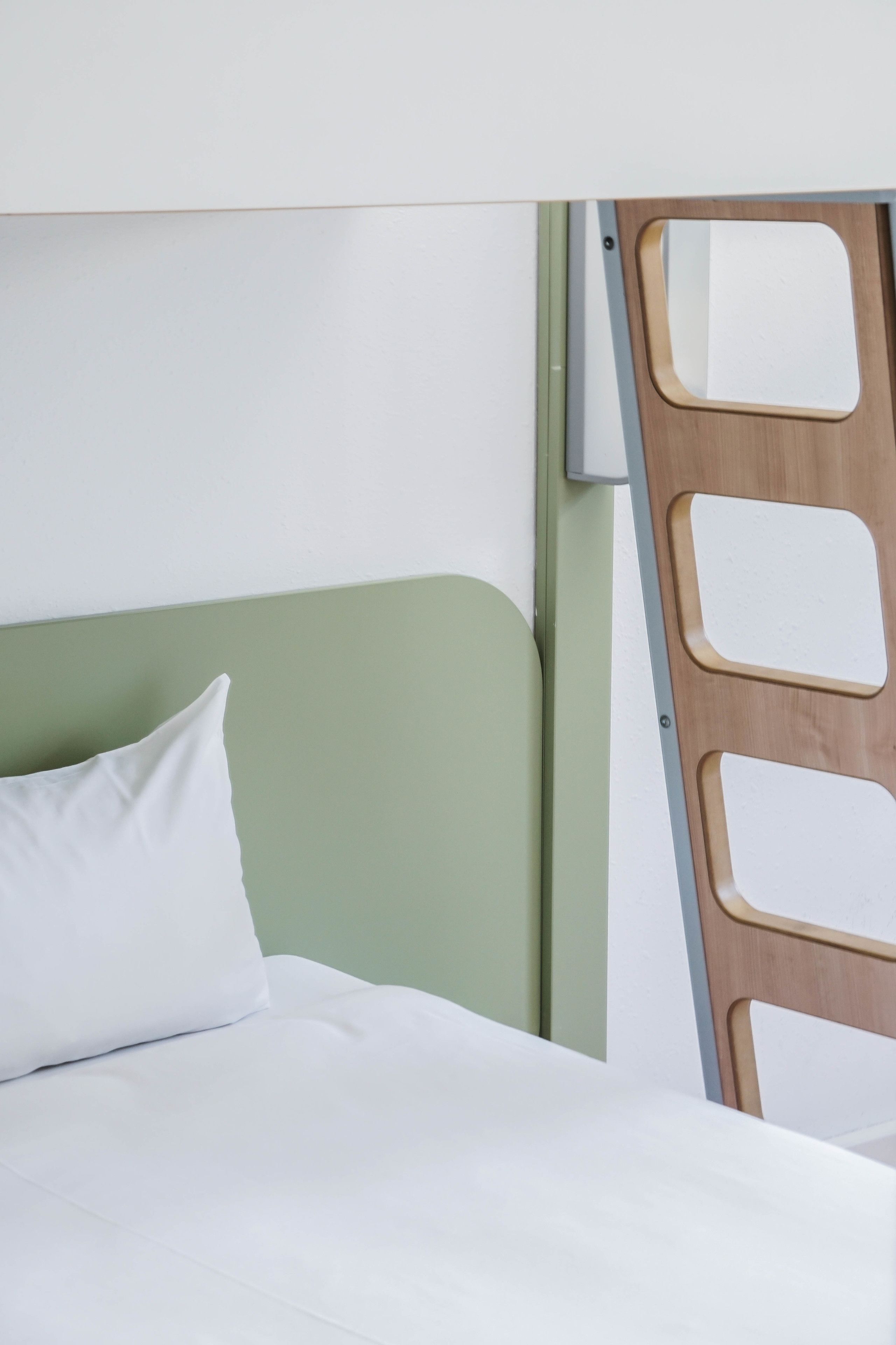 A bed with a green headboard and white bedding, a wooden ladder leaning against the wall, and a small white object on the wall.